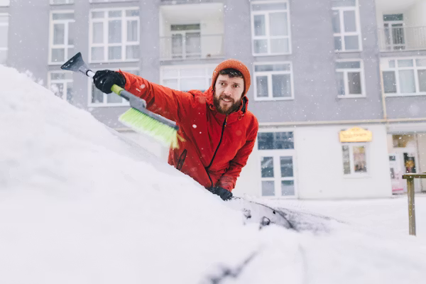 Mies harjaa lunta auton päältä talvella